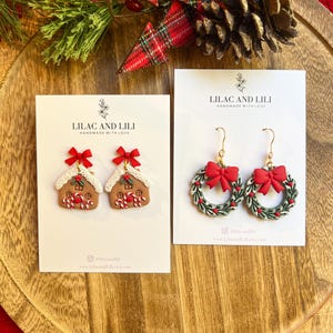 May include: Two pairs of Christmas-themed earrings on white cards. One pair features gingerbread house designs with red bows, the other features green wreath designs with red bows. The cards read "LILAC AND LILI" and "HANDMADE WITH LOVE".