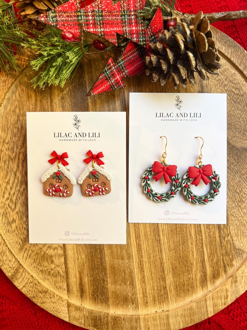 May include: Two pairs of Christmas-themed earrings on white cards. One pair features gingerbread house designs with red bows, the other features green wreath designs with red bows. The cards read "LILAC AND LILI" and "HANDMADE WITH LOVE".