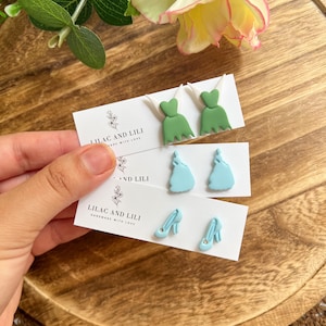 May include: Handmade earrings in the image. The earrings are in the shape of a green dress, a light blue dress, and light blue high heels. The earrings are displayed on white cards with the text "LILAC AND LILI HANDMADE WITH LOVE".