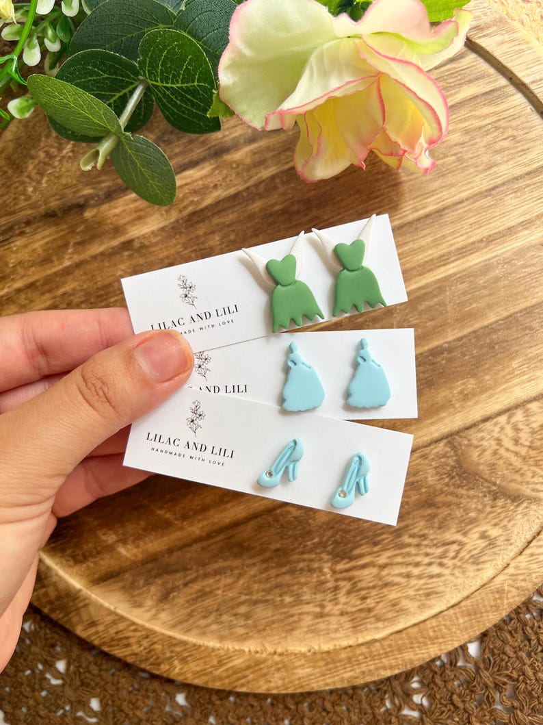 May include: Handmade earrings in the image. The earrings are in the shape of a green dress, a light blue dress, and light blue high heels. The earrings are displayed on white cards with the text "LILAC AND LILI HANDMADE WITH LOVE".