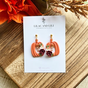 May include: A pair of orange pumpkin-shaped earrings with floral accents. The earrings are handmade and feature a small white flower and a burgundy flower on each pumpkin. The earrings are on a white card with the text "Lilac and Lili Handmade with Love" and the social media handle "@lilacandili" and website "www.lilacandili.etsy.com".