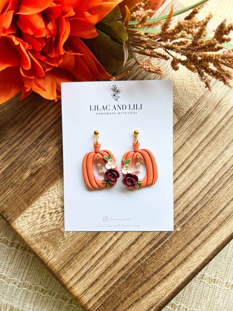 May include: A pair of orange pumpkin-shaped earrings with floral accents. The earrings are handmade and feature a small white flower and a burgundy flower on each pumpkin. The earrings are on a white card with the text "Lilac and Lili Handmade with Love" and the social media handle "@lilacandili" and website "www.lilacandili.etsy.com".