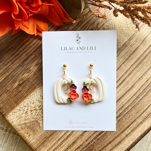 May include: White pumpkin-shaped earrings with a gold hook and a floral design. The flowers are orange, red, and burgundy. The earrings are handmade and say "Lilac and Lili Handmade with Love" on the card.