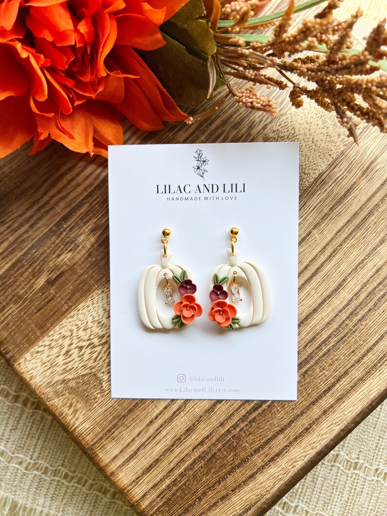 May include: White pumpkin-shaped earrings with a gold hook and a floral design. The flowers are orange, red, and burgundy. The earrings are handmade and say "Lilac and Lili Handmade with Love" on the card.