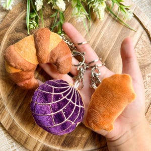 May include: A selection of keyrings, including a croissant, a purple globe with white stitching, and an orange pastry. The keyrings are attached to silver metal rings and clasps. The items are displayed on a wooden tray.