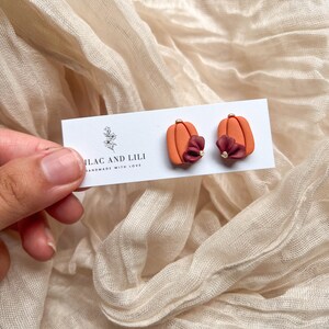 May include: A pair of orange pumpkin-shaped earrings with a small burgundy flower accent and gold accents. The earrings are on a white card with the text "ILAC AND LILI HANDMADE WITH LOVE".