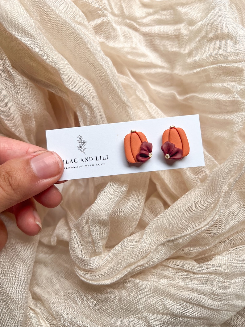 May include: A pair of orange pumpkin-shaped earrings with a small burgundy flower accent and gold accents. The earrings are on a white card with the text "ILAC AND LILI HANDMADE WITH LOVE".