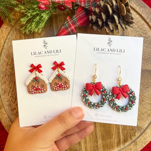 May include: Two pairs of Christmas-themed earrings. One pair features gingerbread house designs with red bows, the other features green wreaths with red bows. Both pairs are on white cards with the text "LILAC AND LILI HANDMADE WITH LOVE."