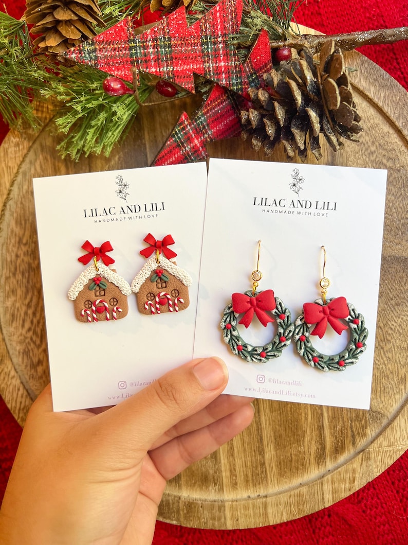 May include: Two pairs of Christmas-themed earrings. One pair features gingerbread house designs with red bows, the other features green wreaths with red bows. Both pairs are on white cards with the text "LILAC AND LILI HANDMADE WITH LOVE."
