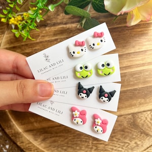 May include: Four pairs of handmade stud earrings featuring cartoon characters. The earrings include designs of a white cat with a pink bow, a green frog, a black cat, and a pink character with a flower. The cards read "LILAC AND LILI HANDMADE WITH LOVE."