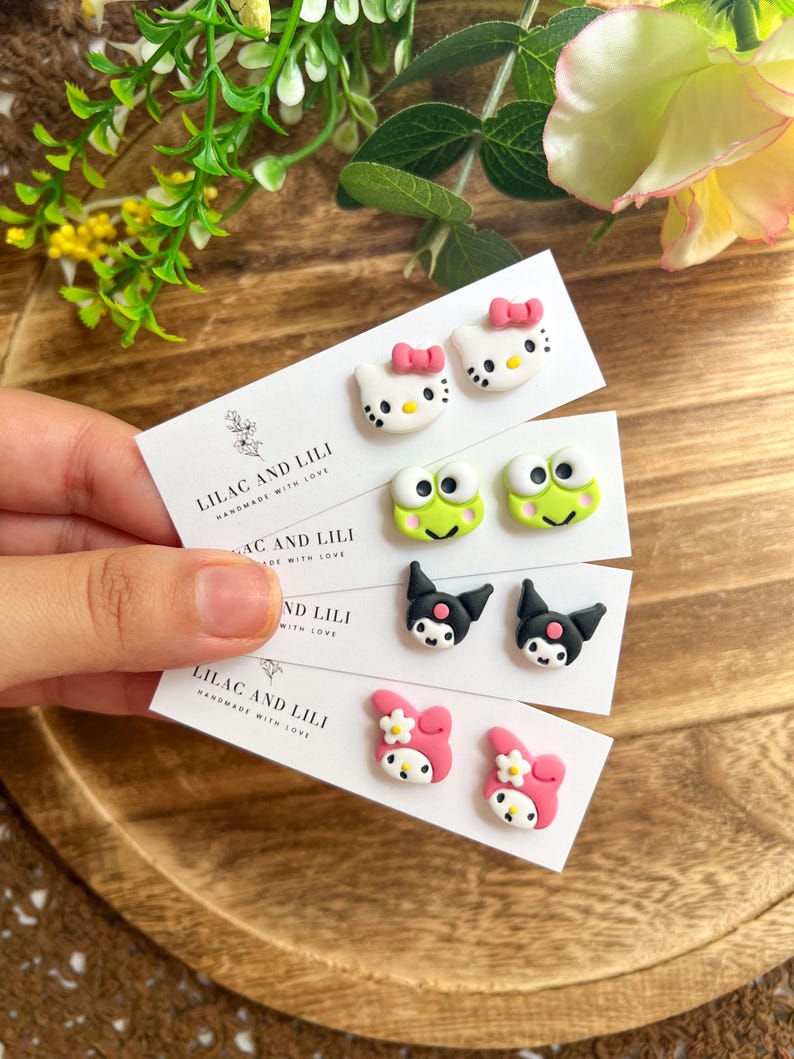 May include: Four pairs of handmade stud earrings featuring cartoon characters. The earrings include designs of a white cat with a pink bow, a green frog, a black cat, and a pink character with a flower. The cards read "LILAC AND LILI HANDMADE WITH LOVE."