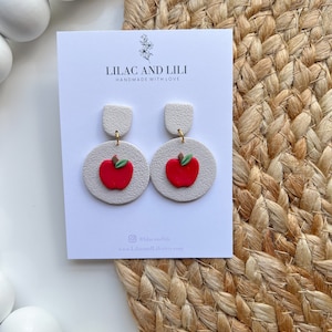 May include: White and red apple-shaped earrings with a gold accent. The earrings are handmade and have the text "Lilac and Lili Handmade with Love" on the card.