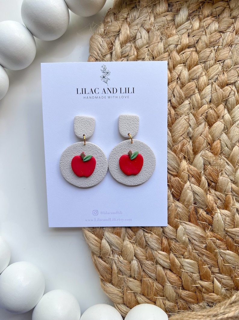 May include: White and red apple-shaped earrings with a gold accent. The earrings are handmade and have the text "Lilac and Lili Handmade with Love" on the card.