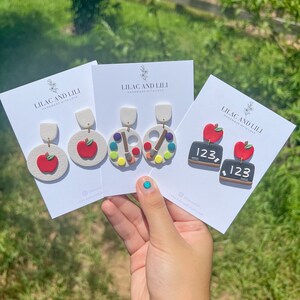 May include: Three pairs of handmade polymer clay earrings. The first pair features a red apple, the second pair features a colourful paint palette and paintbrush, and the third pair features a small chalkboard with the number 123 written on it.