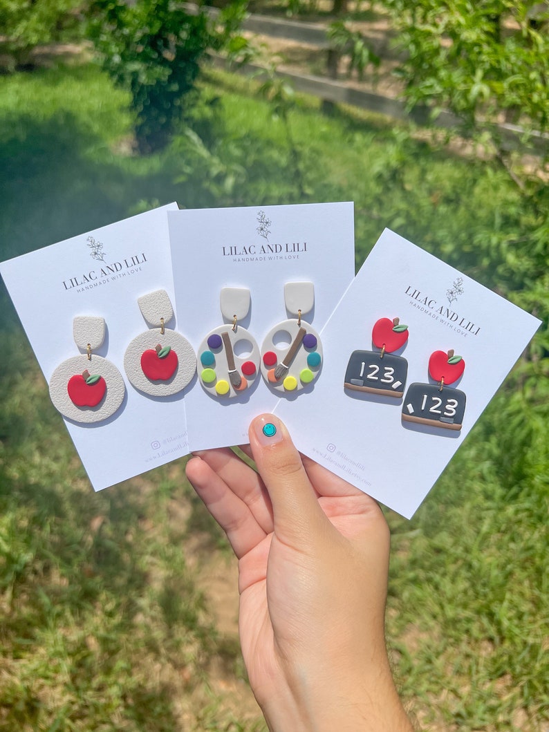 May include: Three pairs of handmade polymer clay earrings. The first pair features a red apple, the second pair features a colourful paint palette and paintbrush, and the third pair features a small chalkboard with the number 123 written on it.