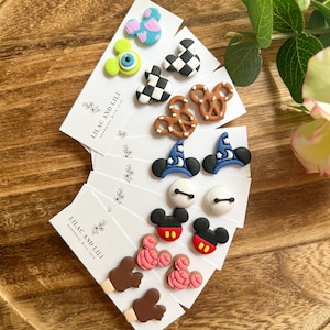 May include: A collection of handmade stud earrings on white cards, featuring various designs like Mickey Mouse, pretzels, and wizard hats. The earrings are in vibrant colors, including red, black, blue, and pink, displayed on a wooden tray.