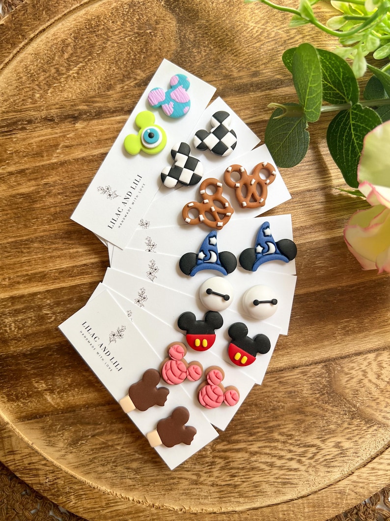 May include: A collection of handmade stud earrings on white cards, featuring various designs like Mickey Mouse, pretzels, and wizard hats. The earrings are in vibrant colors, including red, black, blue, and pink, displayed on a wooden tray.