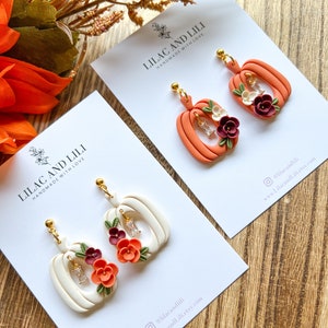 May include: Two pairs of handmade pumpkin earrings with floral accents. The earrings are made of polymer clay and feature a white pumpkin with orange and burgundy flowers and a peach pumpkin with white and burgundy flowers. The earrings are on white cards with the text "Lilac and Lili Handmade with Love" and social media information.
