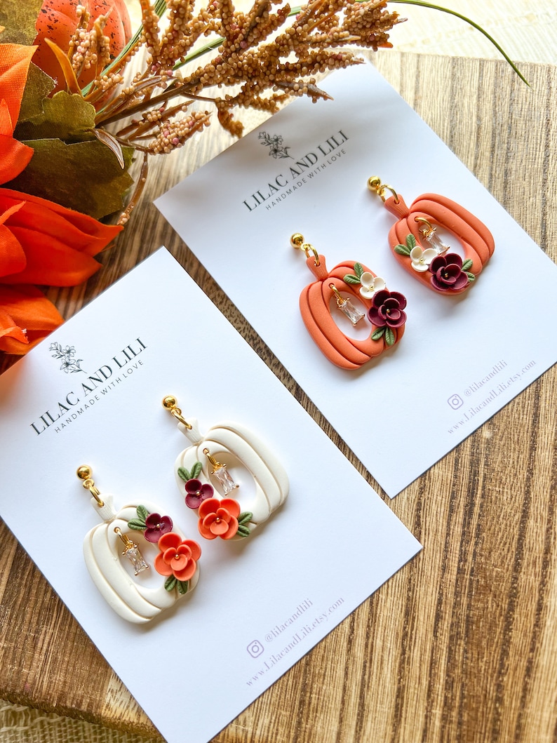 May include: Two pairs of handmade pumpkin earrings with floral accents. The earrings are made of polymer clay and feature a white pumpkin with orange and burgundy flowers and a peach pumpkin with white and burgundy flowers. The earrings are on white cards with the text "Lilac and Lili Handmade with Love" and social media information.