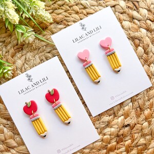 Pu&ograve; includere: Un paio di orecchini fatti a mano con un ciondolo a forma di matita gialla e un ciondolo a forma di cuore rosa o mela rossa. Gli orecchini sono esposti su una carta bianca con il testo "Lilac and Lili Handmade with Love" e informazioni sui social media.