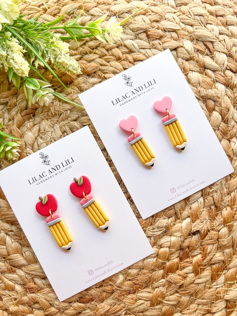 Pu&ograve; includere: Un paio di orecchini fatti a mano con un ciondolo a forma di matita gialla e un ciondolo a forma di cuore rosa o mela rossa. Gli orecchini sono esposti su una carta bianca con il testo "Lilac and Lili Handmade with Love" e informazioni sui social media.