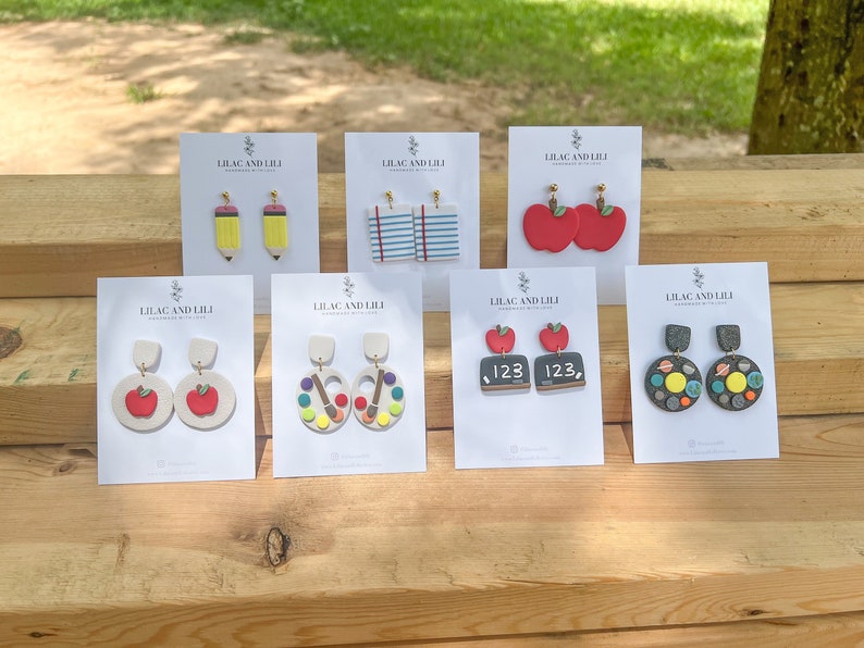 May include: Six cards displaying various pairs of clay earrings. The earrings are themed for teachers and include pencils, paint palettes, apples, chalkboards, and planets. Each card is white with black text that reads "Lilac and Lili Handmade Jewelry".