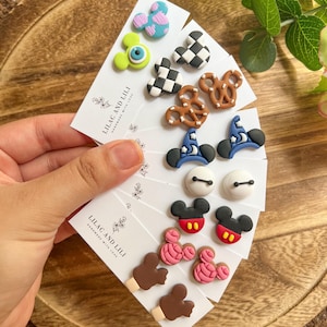 May include: A collection of handmade stud earrings featuring various designs, including cartoon characters, pretzels, and hats. The earrings are displayed on white cards with the text "LILAC AND LILI HANDMADE WITH LOVE". The earrings are in various colors, including blue, pink, black, and white.
