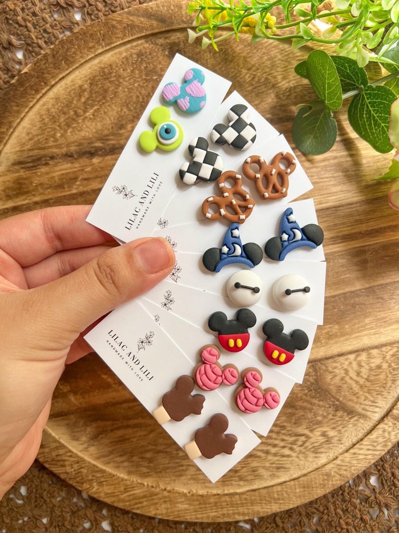 May include: A collection of handmade stud earrings featuring various designs, including cartoon characters, pretzels, and hats. The earrings are displayed on white cards with the text "LILAC AND LILI HANDMADE WITH LOVE". The earrings are in various colors, including blue, pink, black, and white.