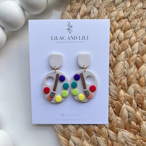 May include: A pair of white and colourful paint palette earrings with a paintbrush detail. The earrings are handmade and feature a white background with a colourful paint palette design. The earrings are perfect for art lovers and anyone who wants to add a touch of colour to their look.