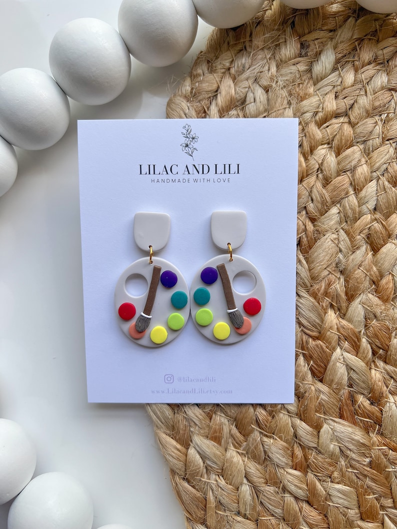 May include: A pair of white and colourful paint palette earrings with a paintbrush detail. The earrings are handmade and feature a white background with a colourful paint palette design. The earrings are perfect for art lovers and anyone who wants to add a touch of colour to their look.