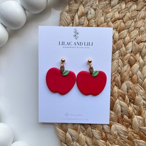 May include: A pair of red apple earrings with green leaves and gold accents. The earrings are handmade and are displayed on a white card with the text "Lilac and Lili Handmade with Love" and the social media handle "@lilacandili".
