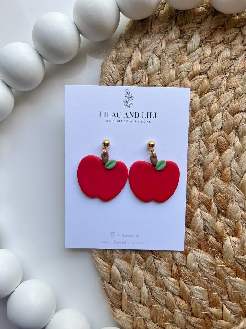 May include: A pair of red apple earrings with green leaves and gold accents. The earrings are handmade and are displayed on a white card with the text "Lilac and Lili Handmade with Love" and the social media handle "@lilacandili".