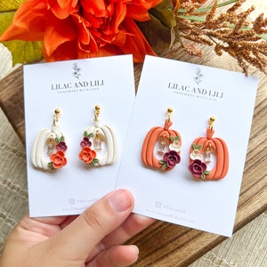May include: Two pairs of pumpkin-shaped earrings with floral accents. The earrings are made of polymer clay and have a white and orange color scheme. The earrings are handmade and have a gold-colored hook.