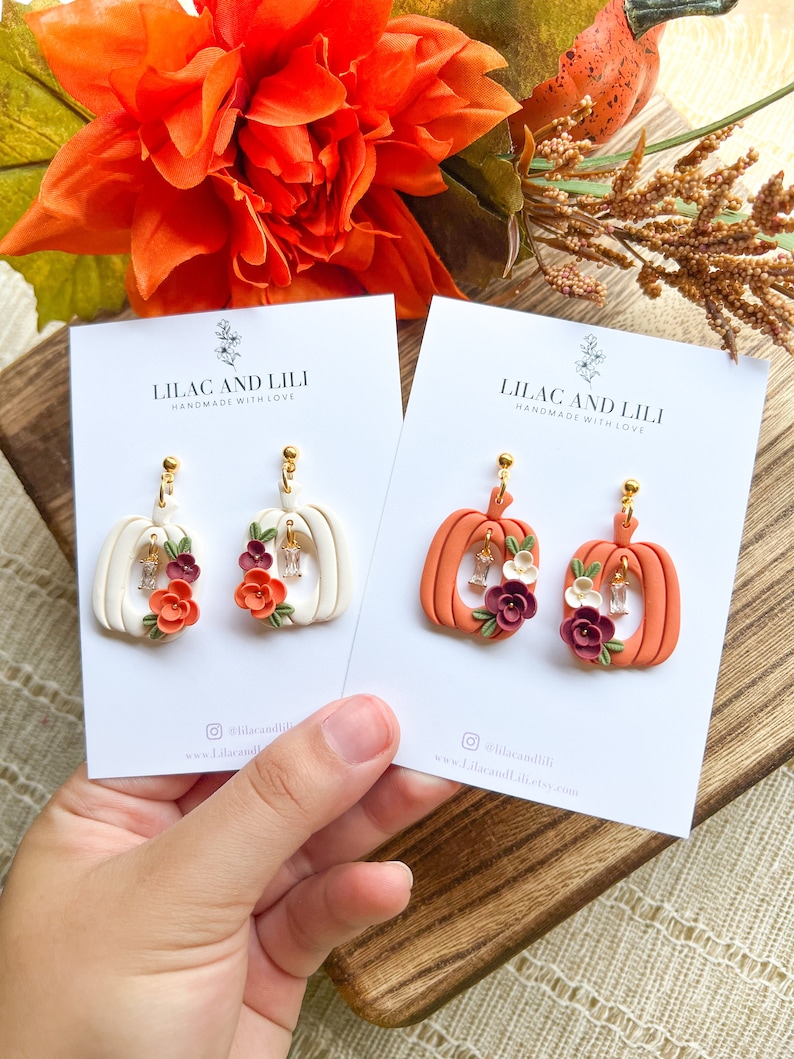 May include: Two pairs of pumpkin-shaped earrings with floral accents. The earrings are made of polymer clay and have a white and orange color scheme. The earrings are handmade and have a gold-colored hook.