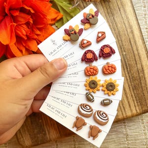 May include: A collection of fall-themed earrings, including turkeys, pumpkin pie slices, sweaters, pumpkins, sunflowers, footballs, cinnamon rolls, and roasted turkeys. The earrings are displayed on white cards with the text "LILAC AND LILI".