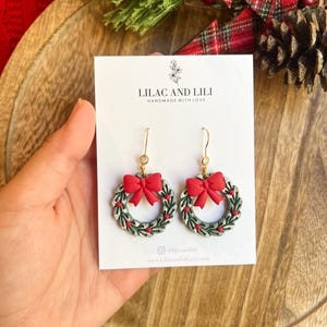 May include: A pair of festive wreath earrings with red bows and small red berry accents. The earrings are gold-toned and feature the text "LILAC AND LILI HANDMADE WITH LOVE". The earrings are displayed on a white card.