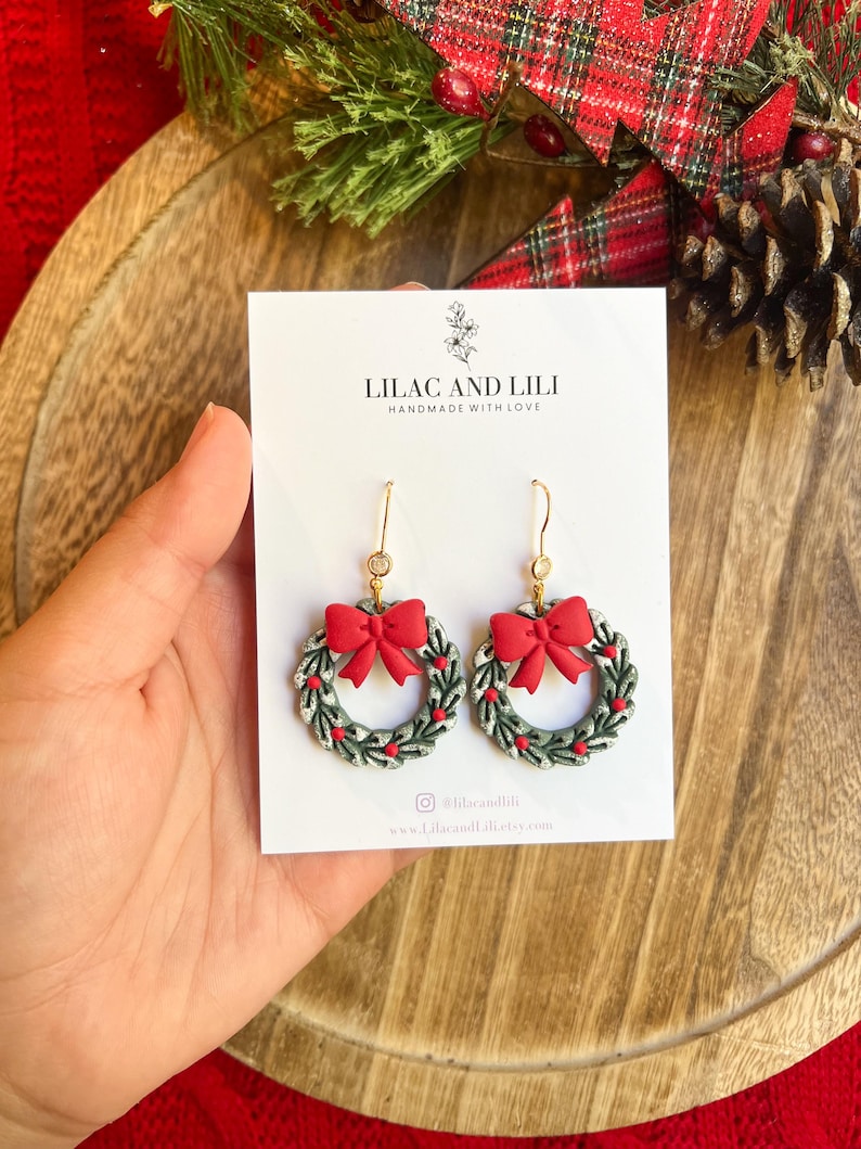 May include: A pair of festive wreath earrings with red bows and small red berry accents. The earrings are gold-toned and feature the text "LILAC AND LILI HANDMADE WITH LOVE". The earrings are displayed on a white card.