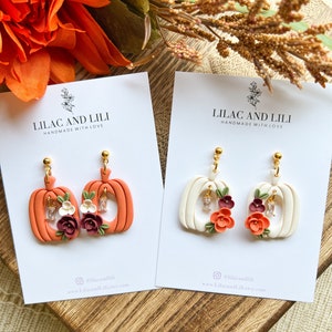 May include: Two pairs of pumpkin-shaped earrings with floral accents. One pair is orange with white accents and the other is white with orange accents. Both pairs feature small rhinestones.