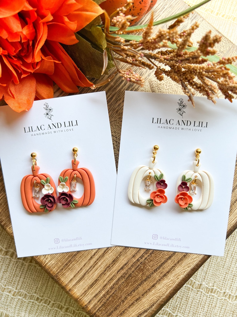 May include: Two pairs of pumpkin-shaped earrings with floral accents. One pair is orange with white accents and the other is white with orange accents. Both pairs feature small rhinestones.