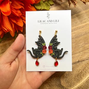 Może przedstawiać: Czarne kolczyki w kształcie kota z kwiatowymi akcentami i czerwonymi kamieniami w kształcie łezki. Kolczyki są na białej karcie z napisem "LILAC AND LILI HANDMADE WITH LOVE".