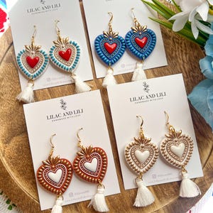 May include: Four pairs of heart-shaped earrings with gold-coloured accents and white tassels. The earrings are in various colours, including red, blue, and brown, with white or red heart centres. Each pair is displayed on a card with the text "LILAC AND LILI".