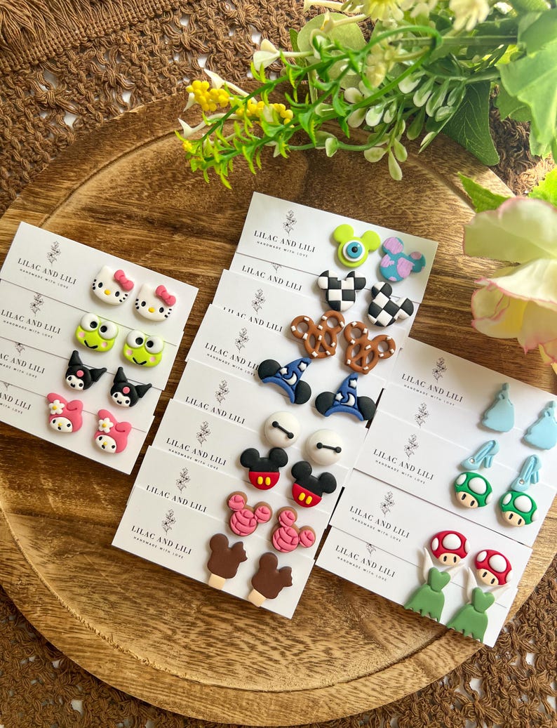 May include: A collection of colorful handmade earrings displayed on white cards, featuring various cartoon characters and food designs. The cards are arranged on a wooden tray, with the text "LILAC AND LILI" visible on each card.