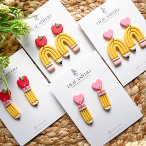 May include: Four pairs of handmade polymer clay earrings. Each pair features a yellow pencil with a pink eraser and a pink heart or red apple charm. The earrings are displayed on white cards with the text "Lilac and Lili Handmade with Love".