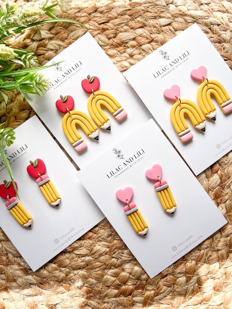 Pu&ograve; includere: Quattro paia di orecchini fatti a mano in pasta polimerica. Ogni paio presenta una matita gialla con una gomma rosa e un ciondolo a forma di cuore rosa o mela rossa. Gli orecchini sono esposti su cartoncini bianchi con il testo "Lilac and Lili Handmade with Love".