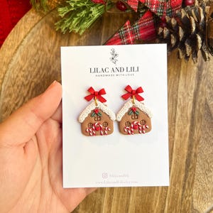 May include: A pair of gingerbread house earrings with red bows, candy cane accents, and white icing details. The earrings are displayed on a white card with the text "LILAC AND LILI HANDMADE WITH LOVE".