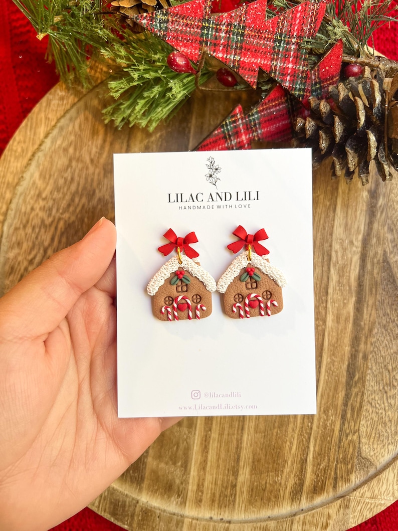 May include: A pair of gingerbread house earrings with red bows, candy cane accents, and white icing details. The earrings are displayed on a white card with the text "LILAC AND LILI HANDMADE WITH LOVE".