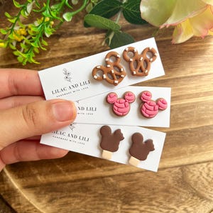 May include: Three pairs of handmade earrings on white cards. The earrings feature pretzel, pink mouse head, and ice cream designs. The cards read "LILAC AND LILI HANDMADE WITH LOVE". The earrings are displayed on a wooden tray.