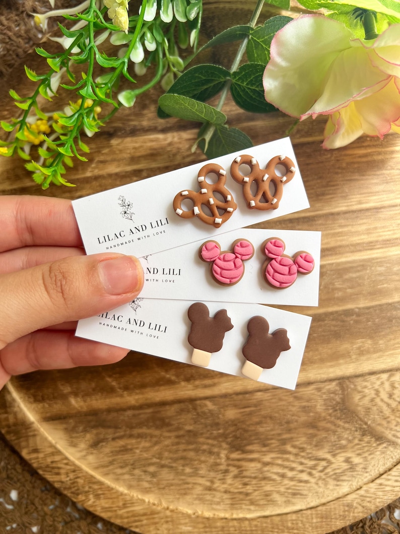 May include: Three pairs of handmade earrings on white cards. The earrings feature pretzel, pink mouse head, and ice cream designs. The cards read "LILAC AND LILI HANDMADE WITH LOVE". The earrings are displayed on a wooden tray.