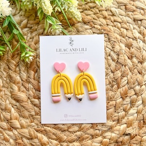 Pu&ograve; includere: Un paio di orecchini a forma di matita gialla e rosa con un ciondolo a forma di cuore rosa. Gli orecchini sono fatti a mano con amore e sono disponibili per l'acquisto su Etsy. Il testo sulla carta recita "Lilac and Lili Handmade with Love" e "@lilacandili www.lilacandili.etsy.com".