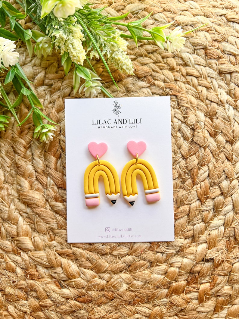 Pu&ograve; includere: Un paio di orecchini a forma di matita gialla e rosa con un ciondolo a forma di cuore rosa. Gli orecchini sono fatti a mano con amore e sono disponibili per l'acquisto su Etsy. Il testo sulla carta recita "Lilac and Lili Handmade with Love" e "@lilacandili www.lilacandili.etsy.com".